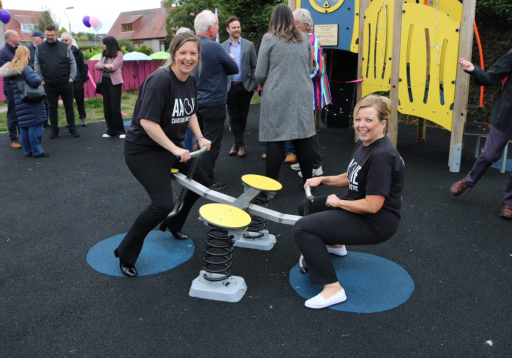 Two women on a seesaw at a playpark, one high in the air, one with feet on the ground