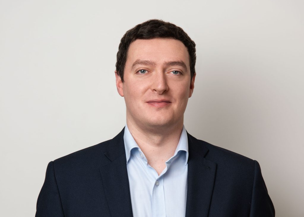 Ani image of Stuart Clark smiling at the camera. He is wearing a black blazer and blue shirt.