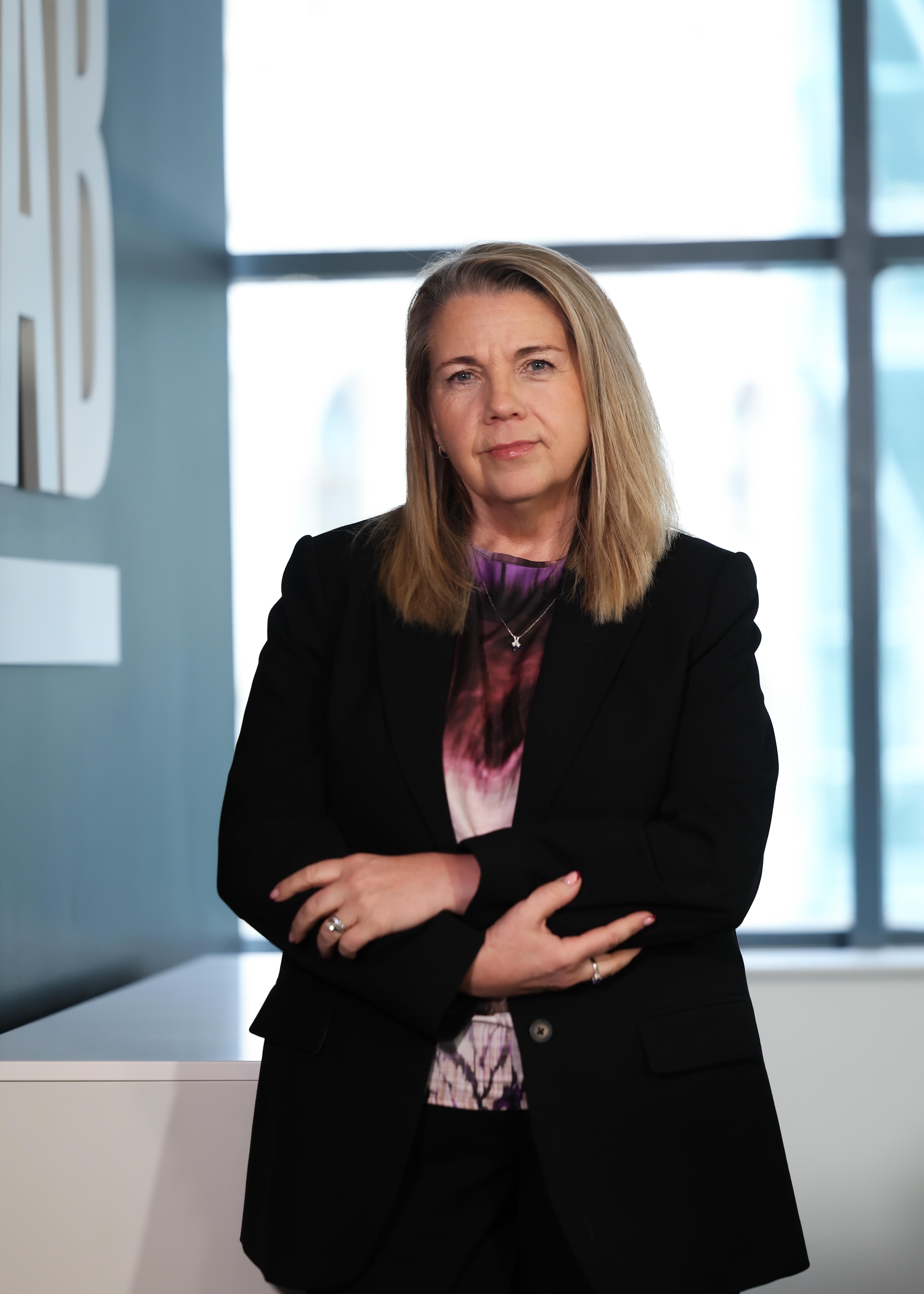 An image of Michelle Hawkins standing in the office wearing a black blazer and purple top. Her arms are clasped together.