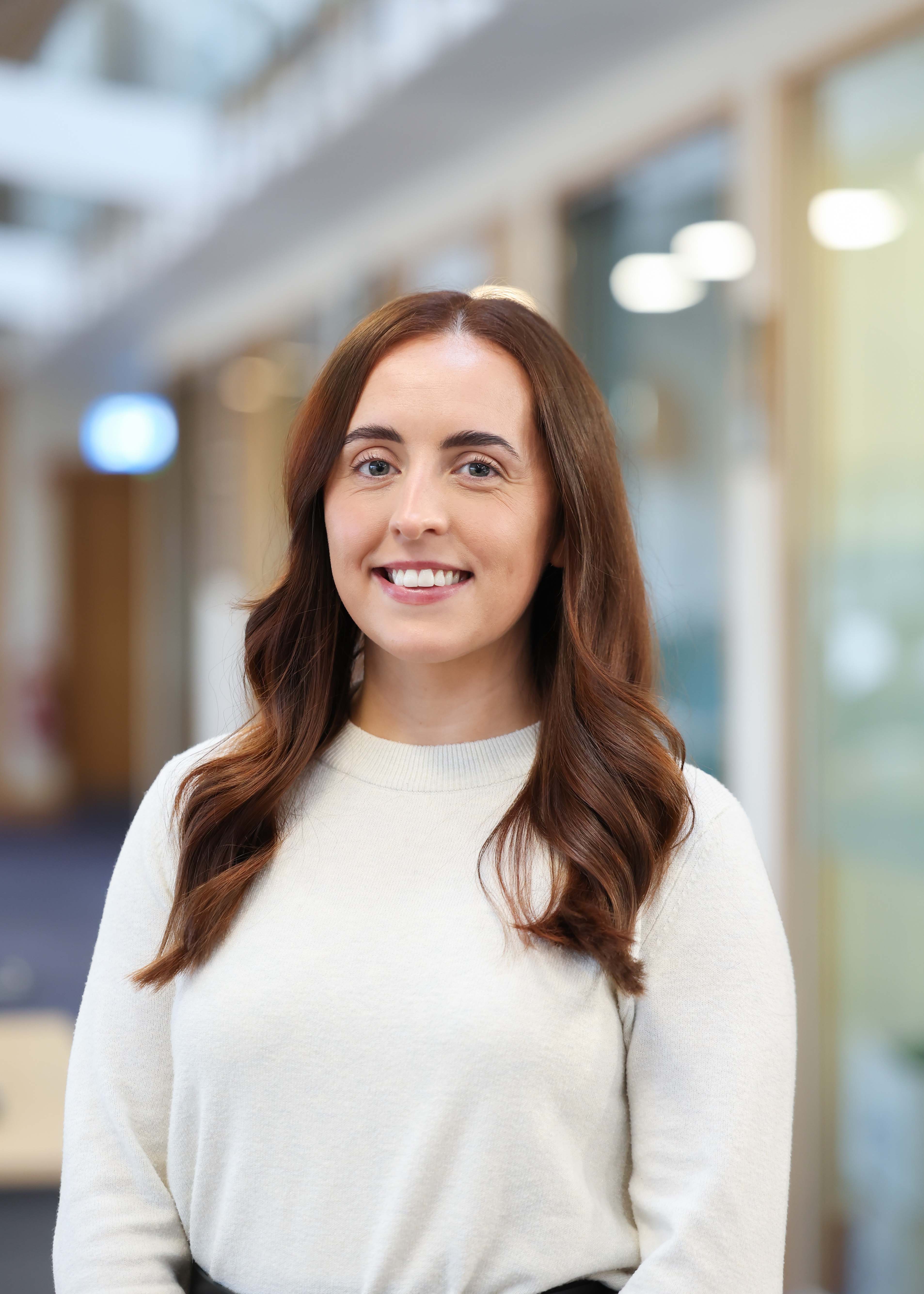 An image of Lauren McMahon standing in the office, wearing a white top and smiling at the camera.
