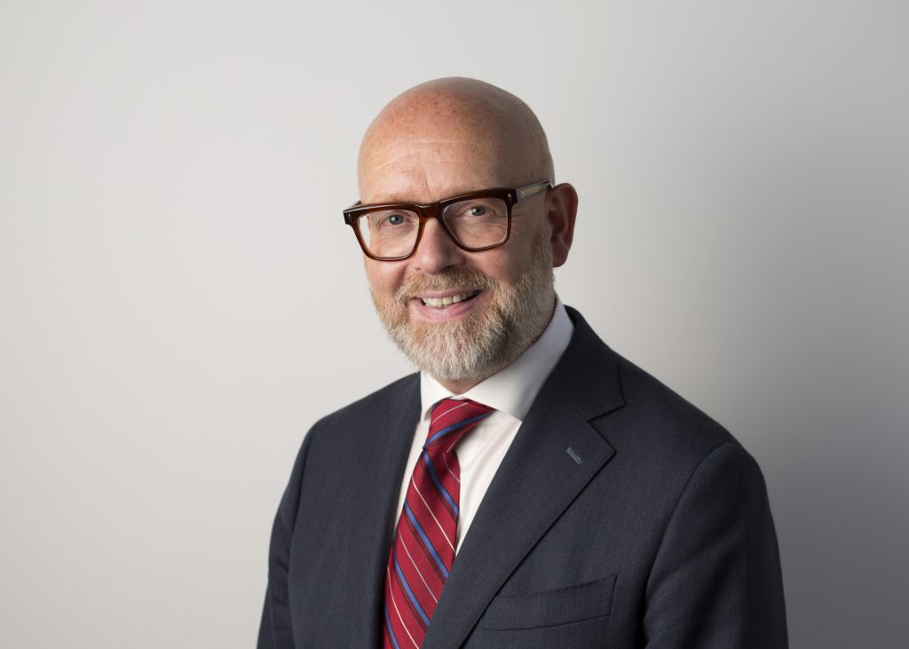 An image of Duncan Glassey wearing a suit and glasses and smiling at the camera.