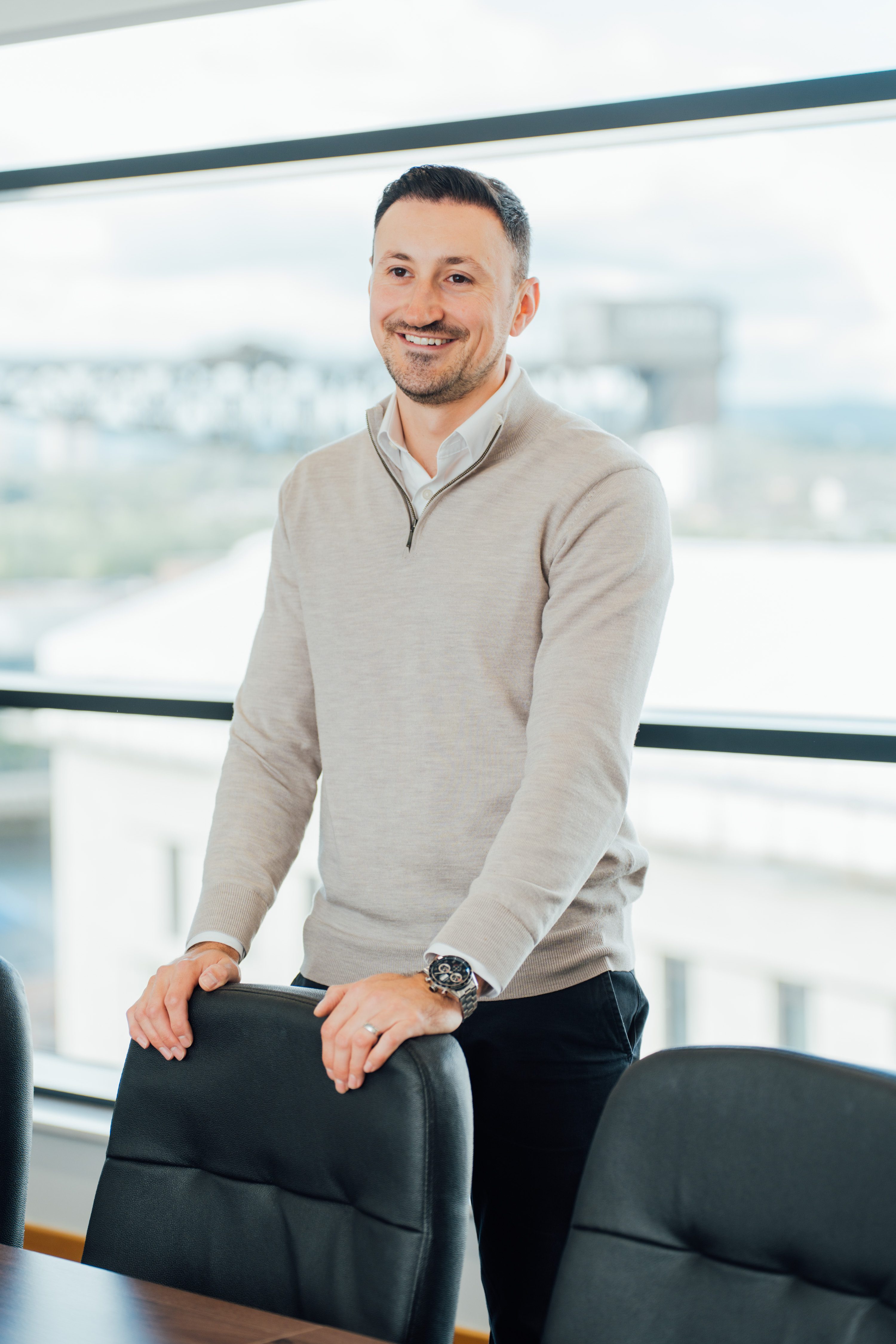 An image of Alistair Stewart wearing a grey jumper and standing in front of a chair.