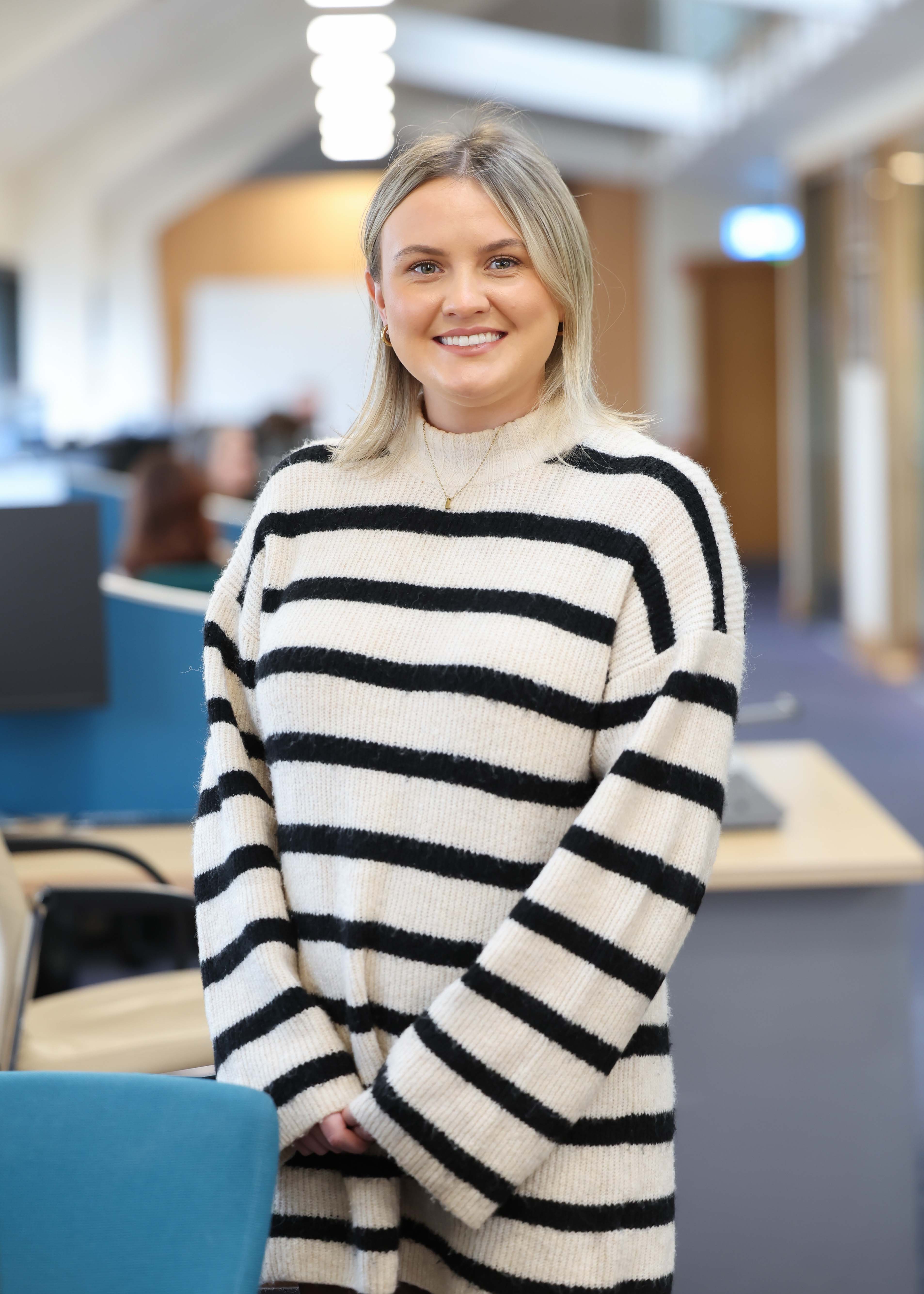 An image of Laura OHagan wearing a striped jumper. She is standing in the office with her hands together.