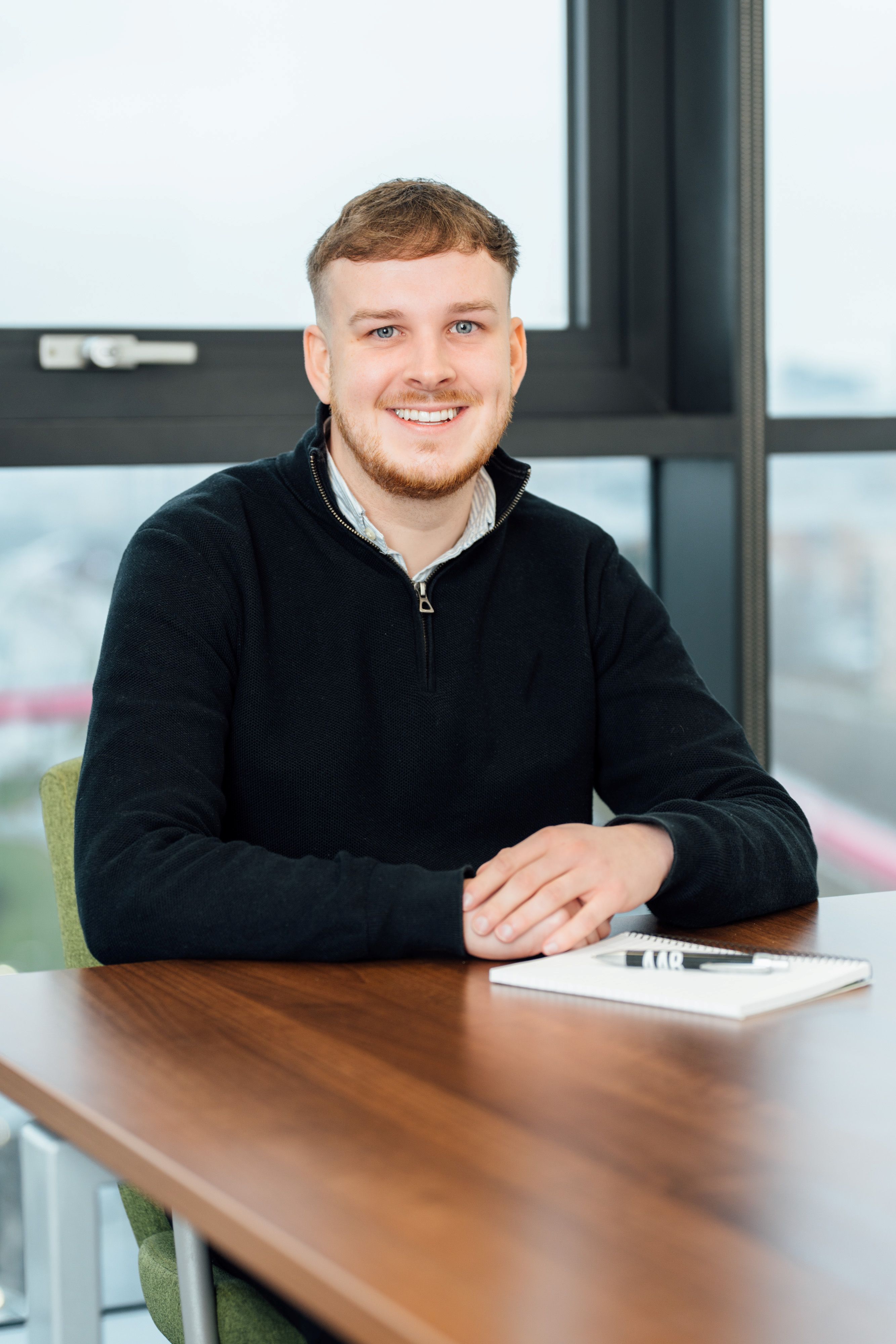 Kyle Sinclair sitting at desk smiling at camera with notebook