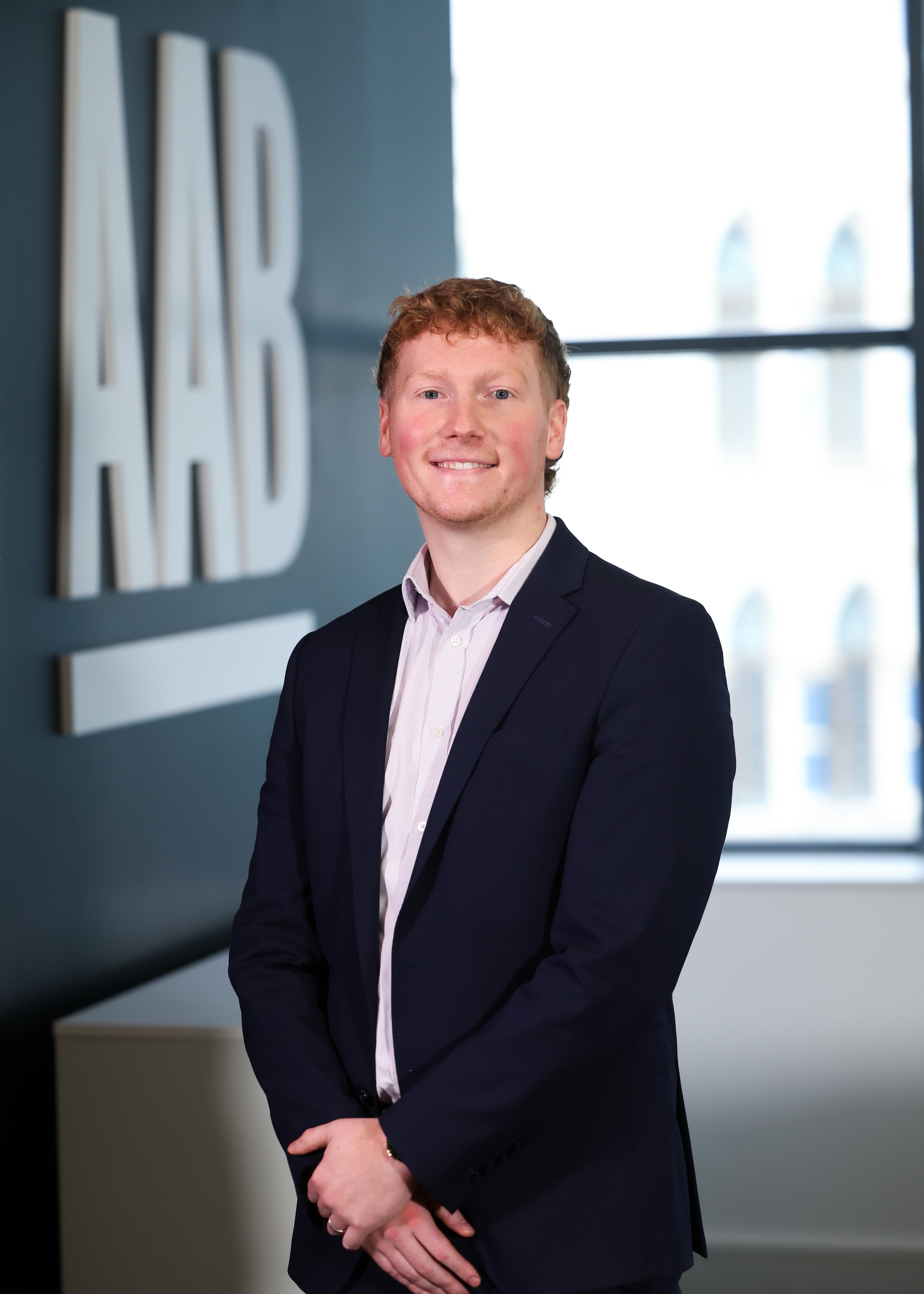 An image of Alexander Hill wearing a black blazer with a pink shirt. He is standing in the office with his hands together,