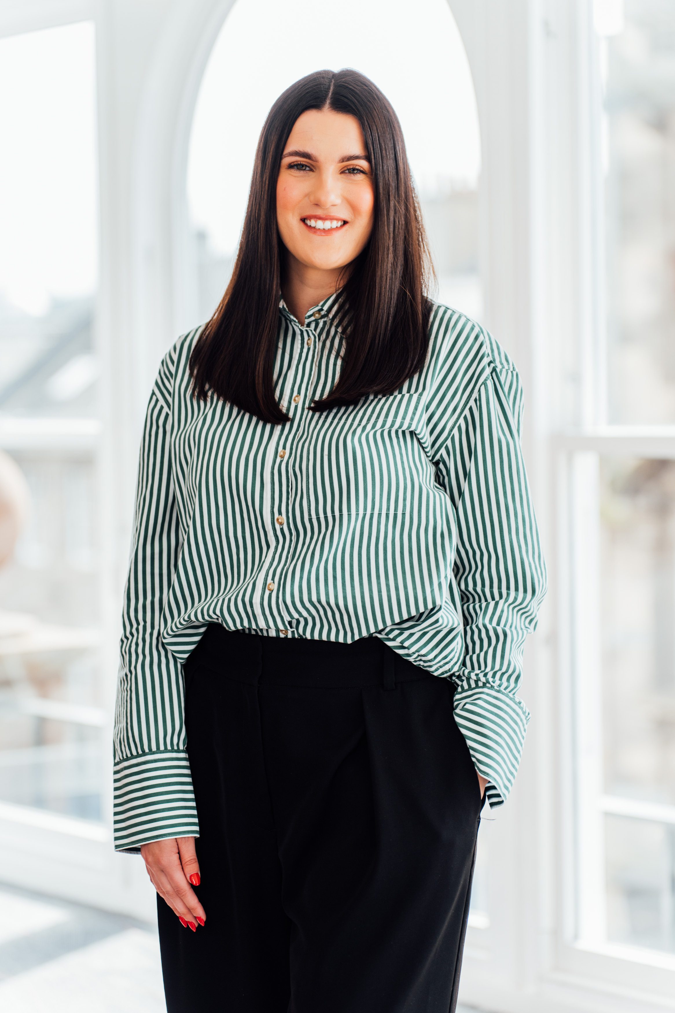 An image of Alix Baird standing by the window with a green shirt and black trousers.
