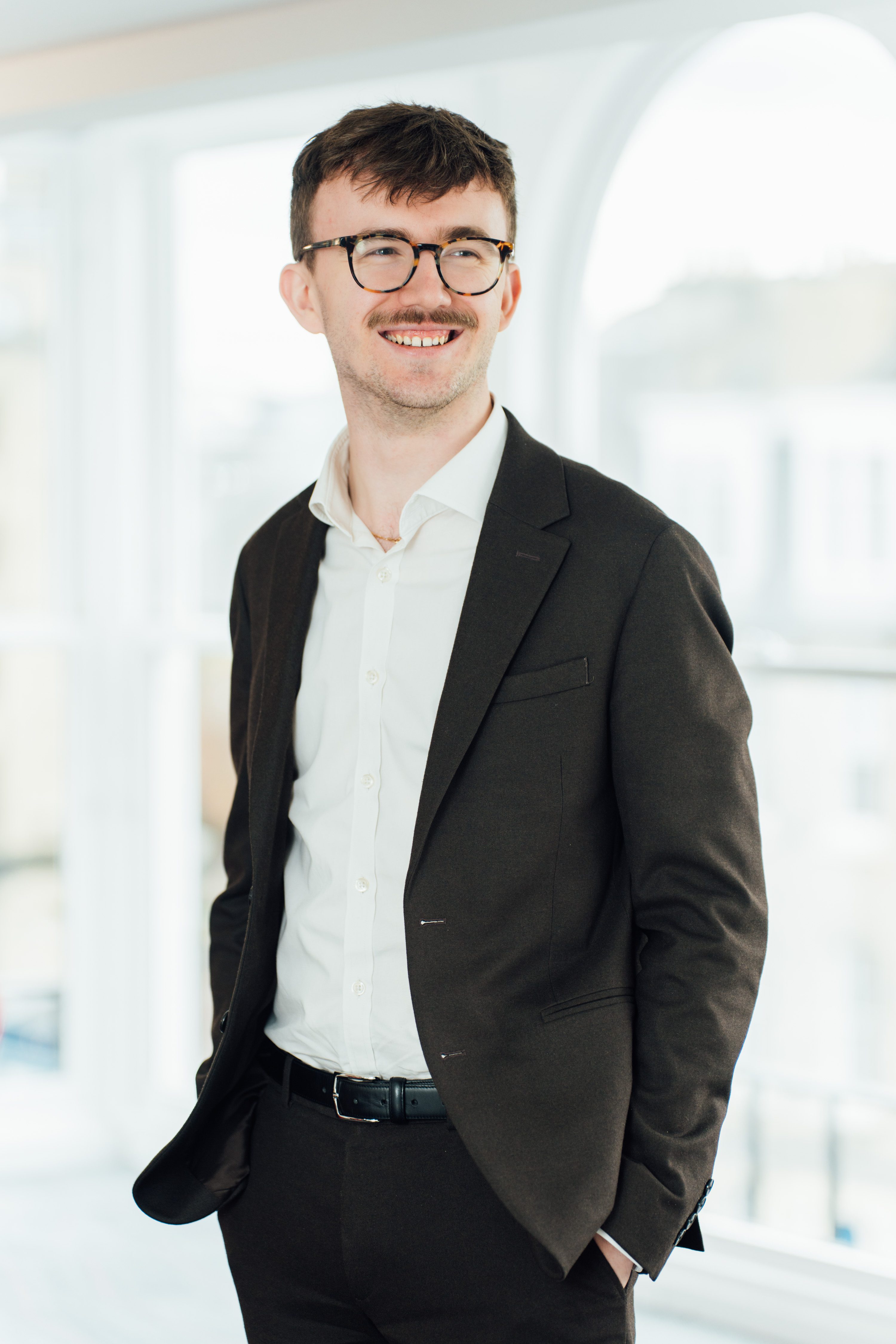 An image of Andrew Philbin wearing a blazer with a white shirt. He is standing smiling with his hands in his pocket.