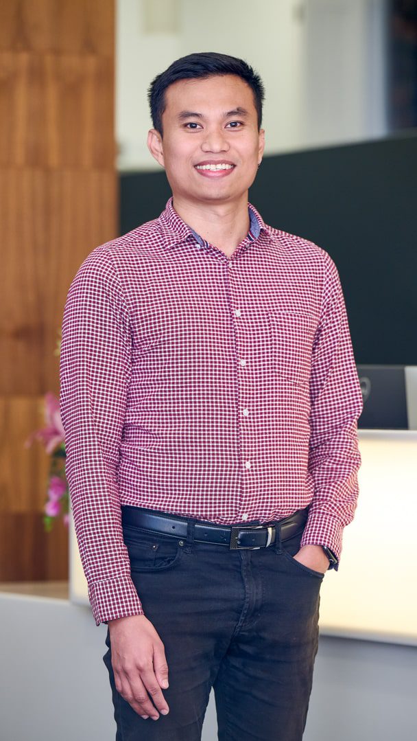 An image of Amiel Malundas standing in the office wearing black trousers and a burgandy shirt. He's smiling at the camera.