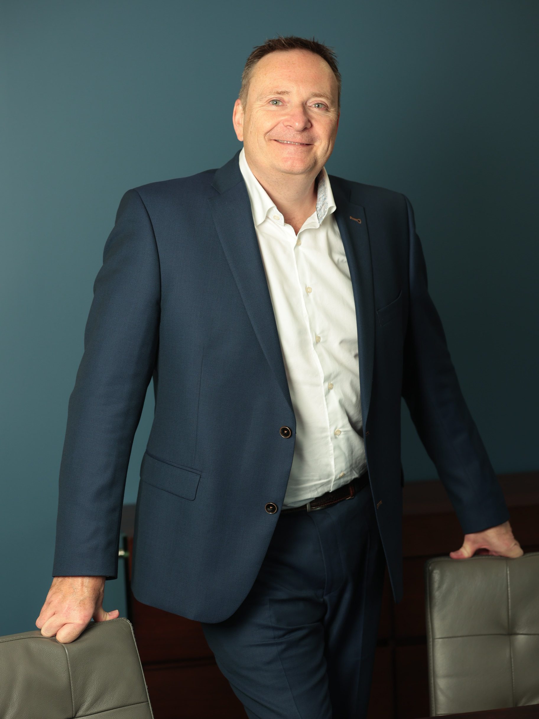 An image of Dante Peters standing next to a chair and wearing a white shirt and navy suit.