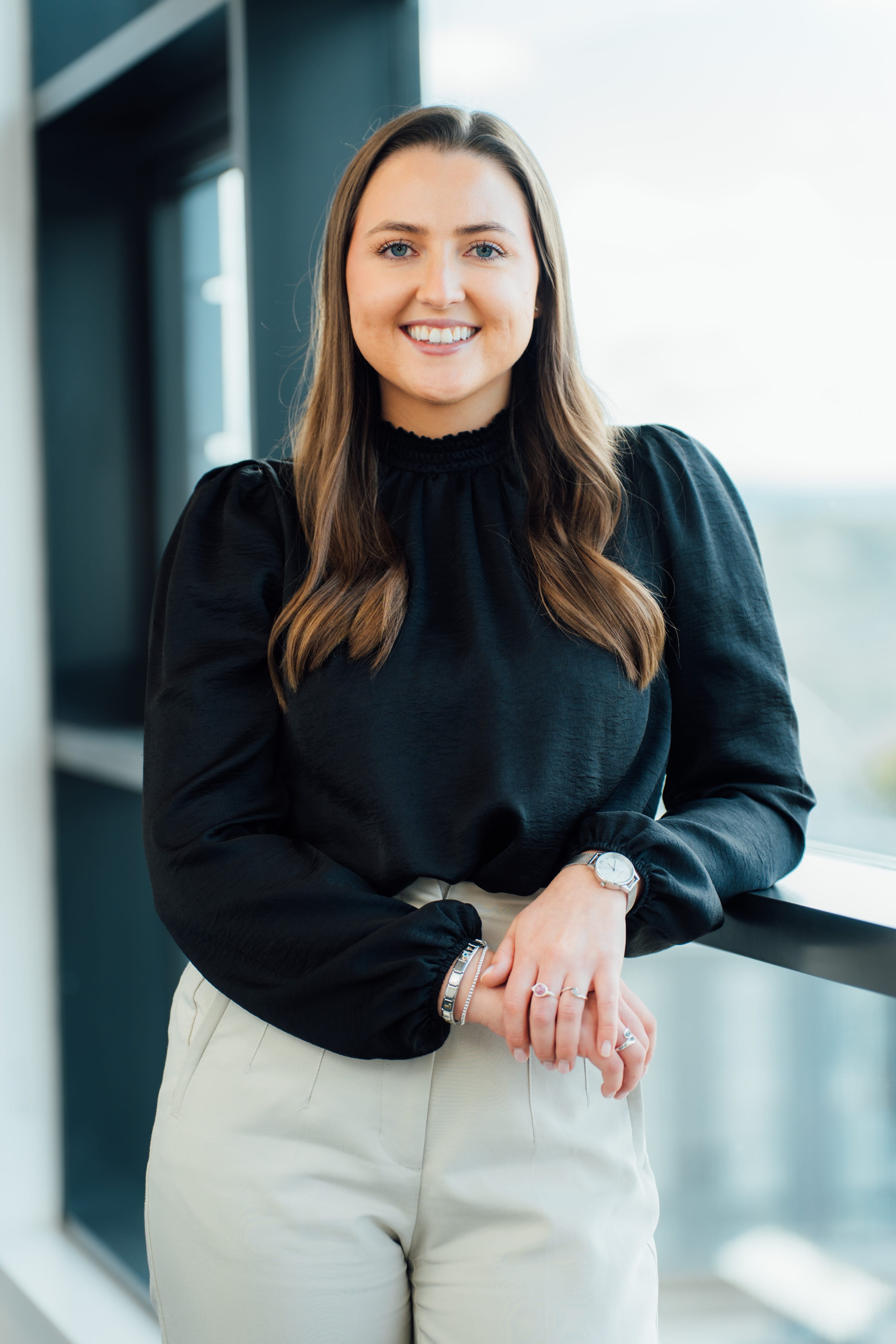 An image of Lori Young standing against a window sill wearing a black top and cream trousers.