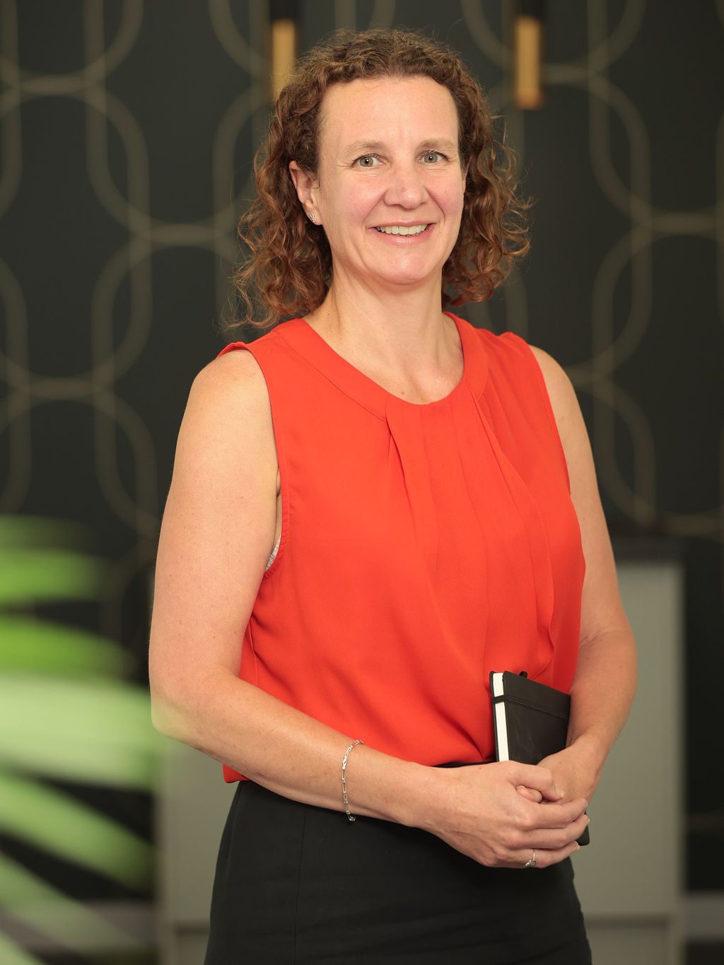 An image of Angela Winton standing in the office and holding a notebook. She is wearing a red top and black trousers.