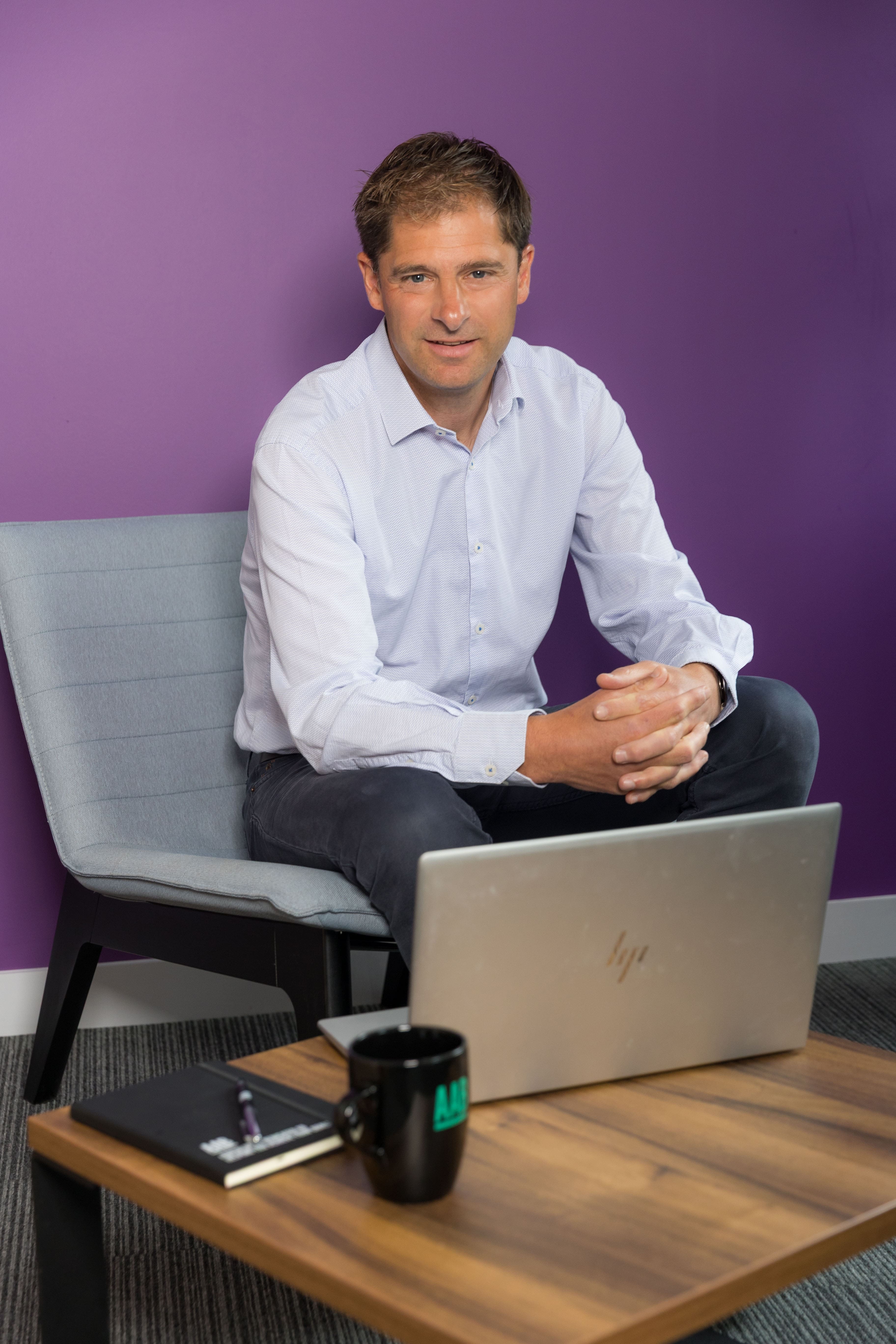 An image of Colin Judd sitting on a chair with his laptop on a desk. There is also a mug and a notebook.