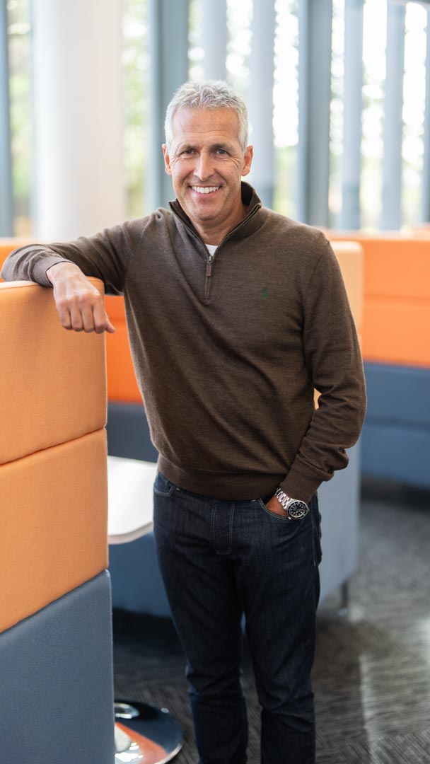 An image of Glenn Brophy standing in the office and leaning on a office chair.