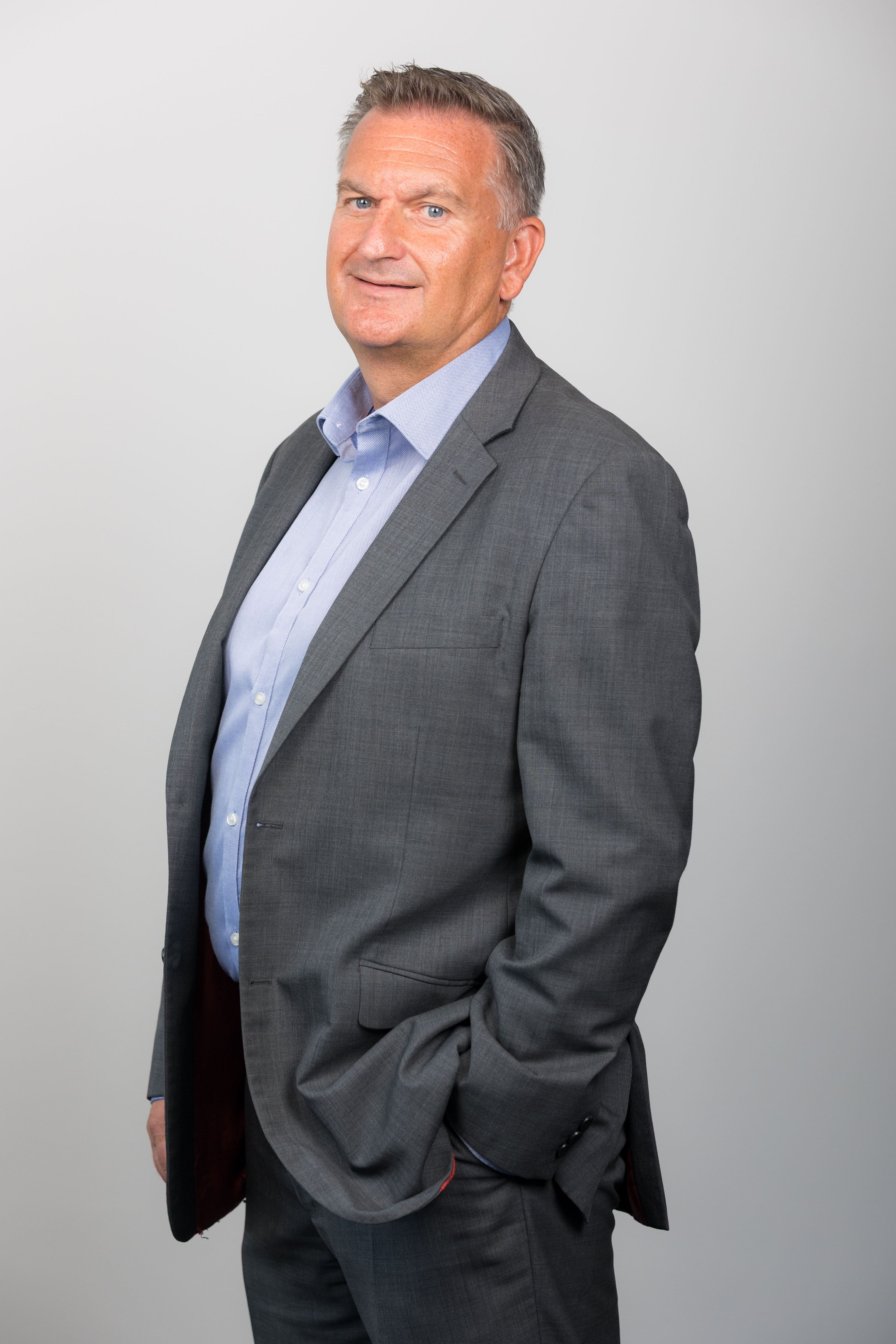 An image of James West standing in front of a grey wall wearing a grey suit jacket and blue shirt.