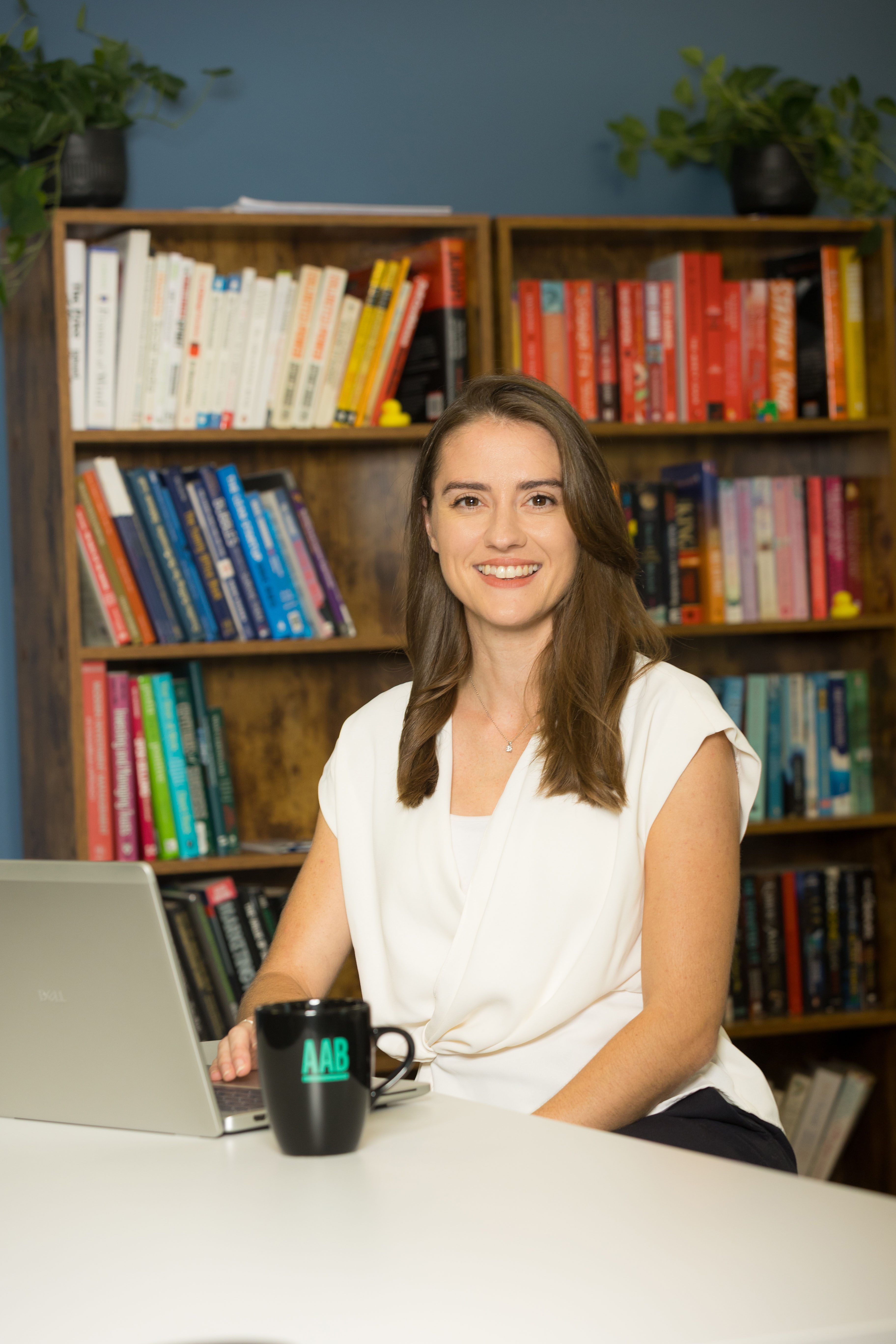 An image of Lizzie Hollamby wearing a white top and sitting at a desk with her AAB branded mug and laptop.