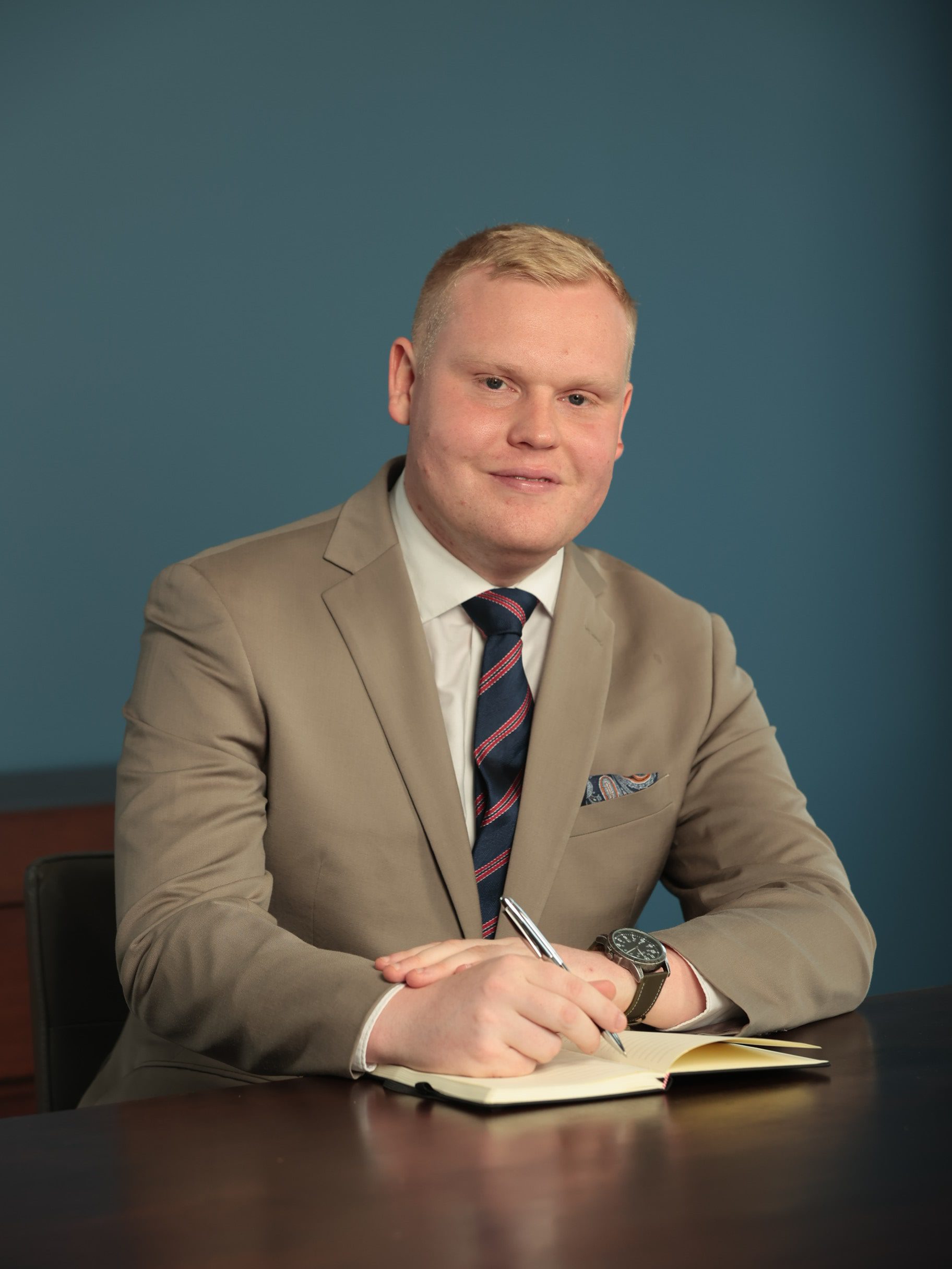 An image of Matthew Knight wearing a beige suit and tie and writing in a notebook.