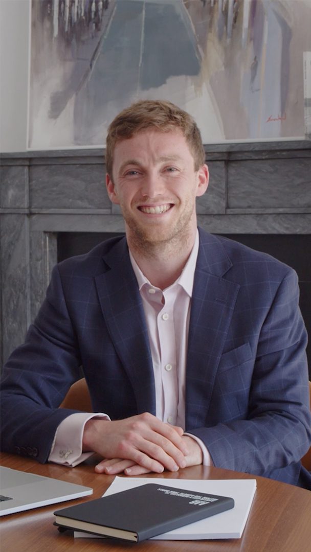 An image of Patrick Christie sitting at a desk with a blazer and shirt.