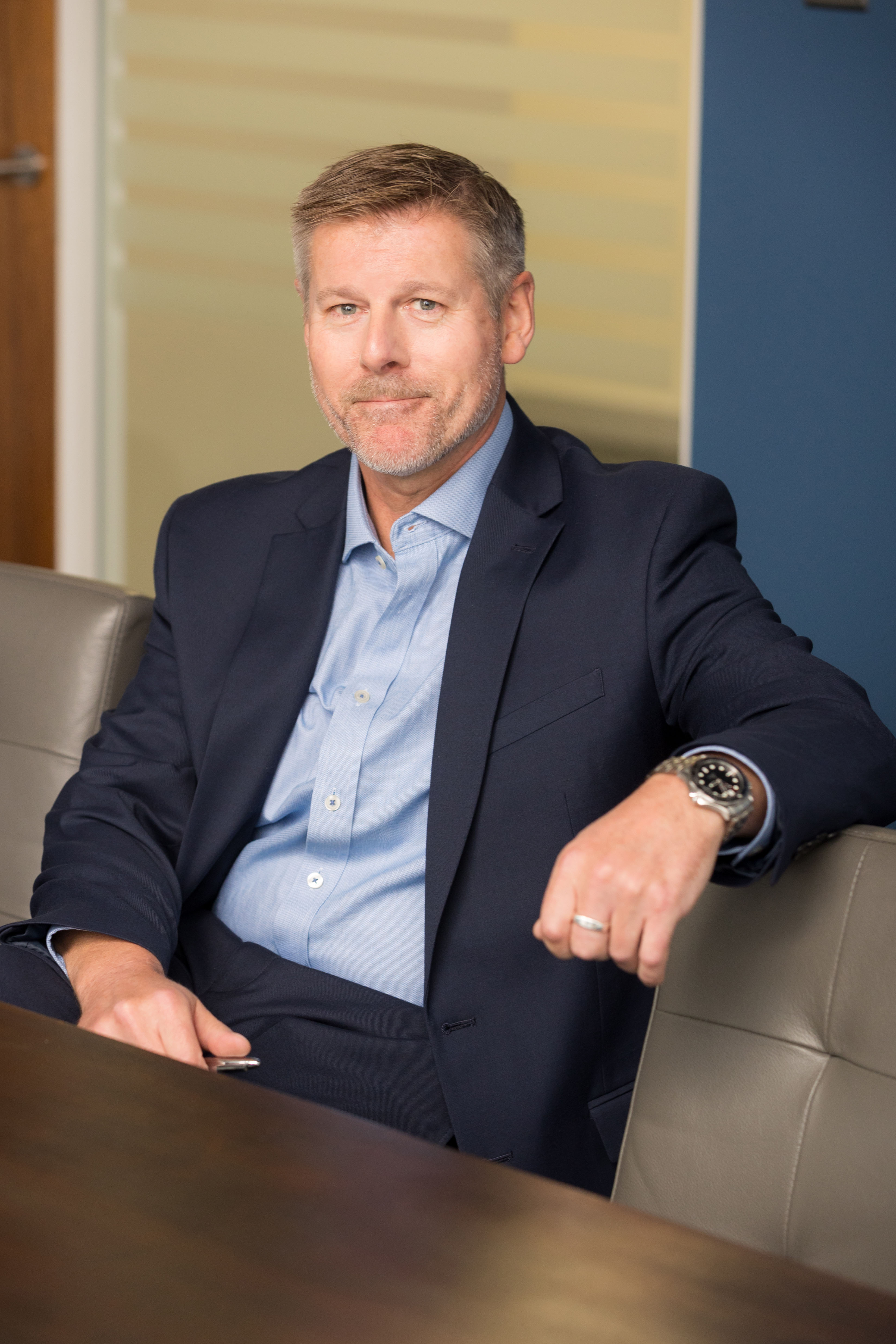An image of Simon Reynolds sitting on a chair at a desk and smiling at the camera.