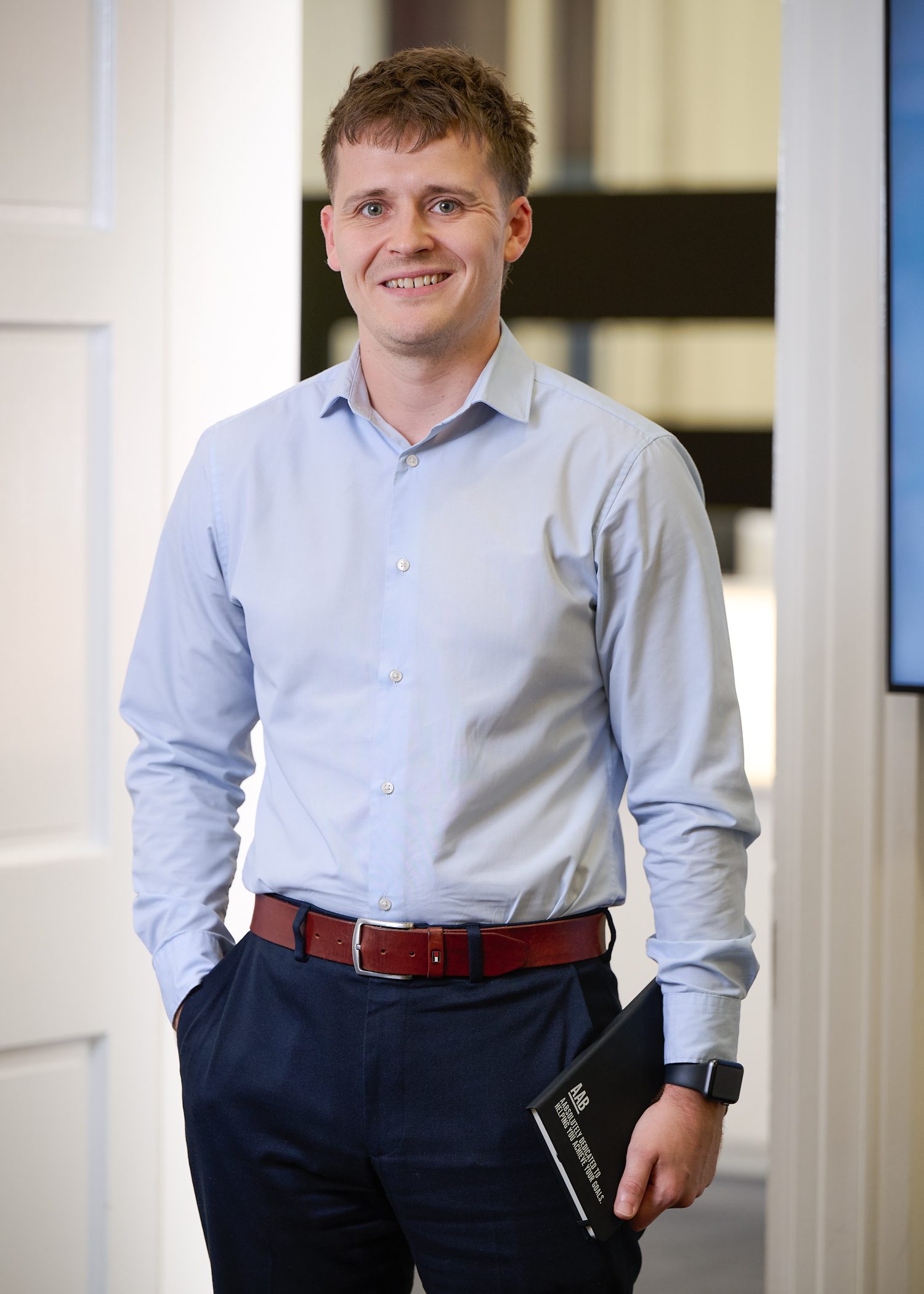 An image of James Taylor standing in the office wearing a blue shirt and black trousers.