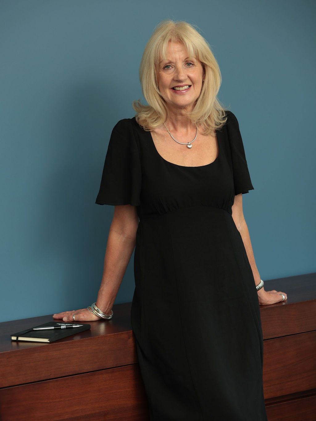 An image of Tracey Thorpe wearing a black dress and leaning on a table. She has blonde hair and is smiling at the camera.