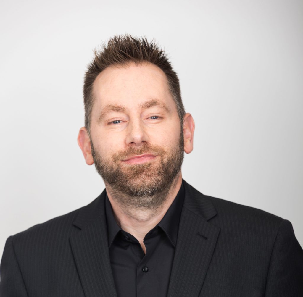 An image of Damien Coughlan wearing a black blazer with a black shirt. He is smiling at the camea