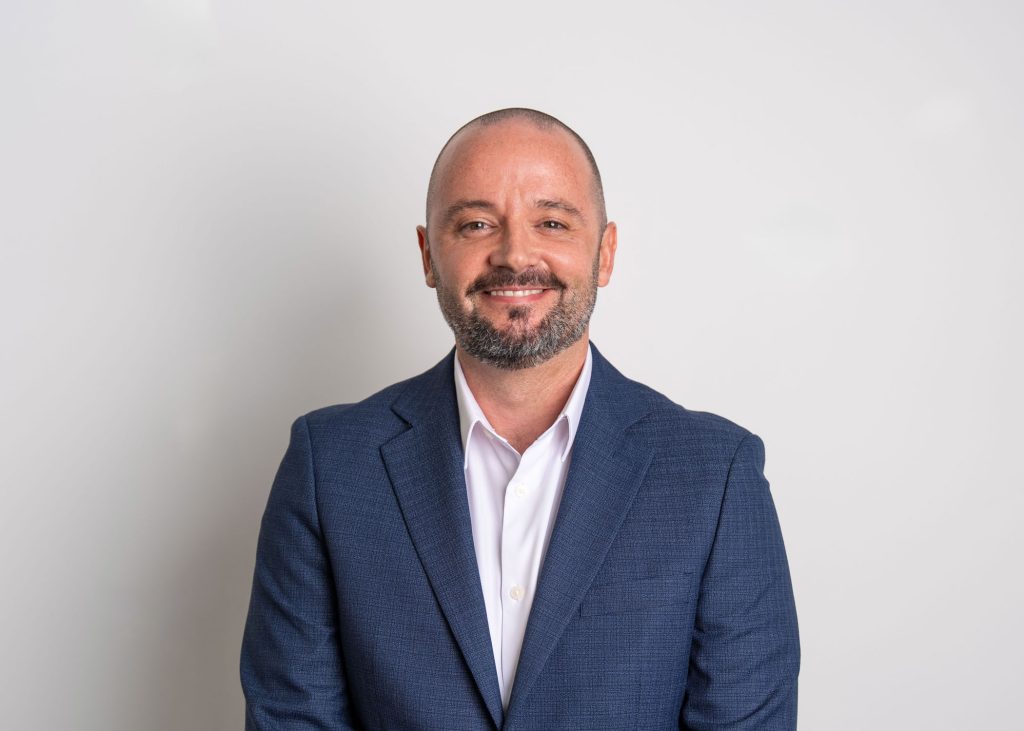 An image of Greg McSneddon wearing a blue blazer with a white shirt. He is smiling at the camera.