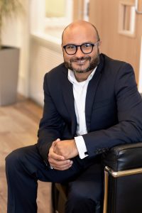 An image of Muhammad Sharif sitting on a chair wearing a black jacket and white shirt.