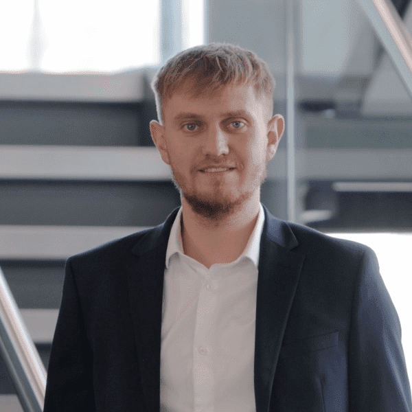 An image of Thomas Booth standing in the office wearing a white shirt and navy suit. He has his hand in his pocket.