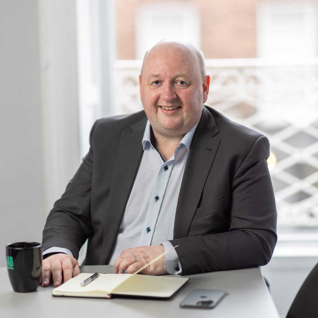 An image of Aidan McCarthy wearing a grey blazer and blue shirt. He is smiling at the camera.