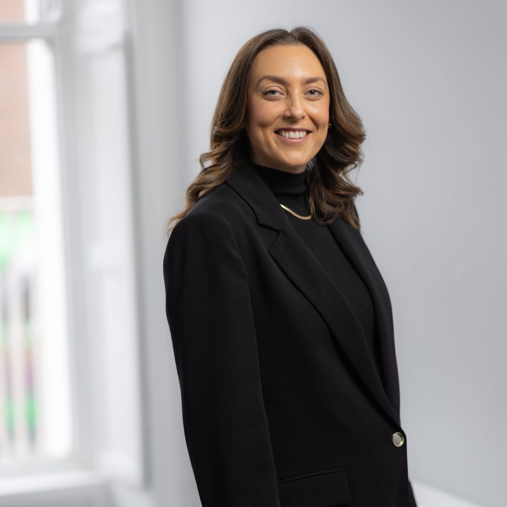 An image of Ciara McDunphy wearing a black blazer and standing next to a desk smiling.