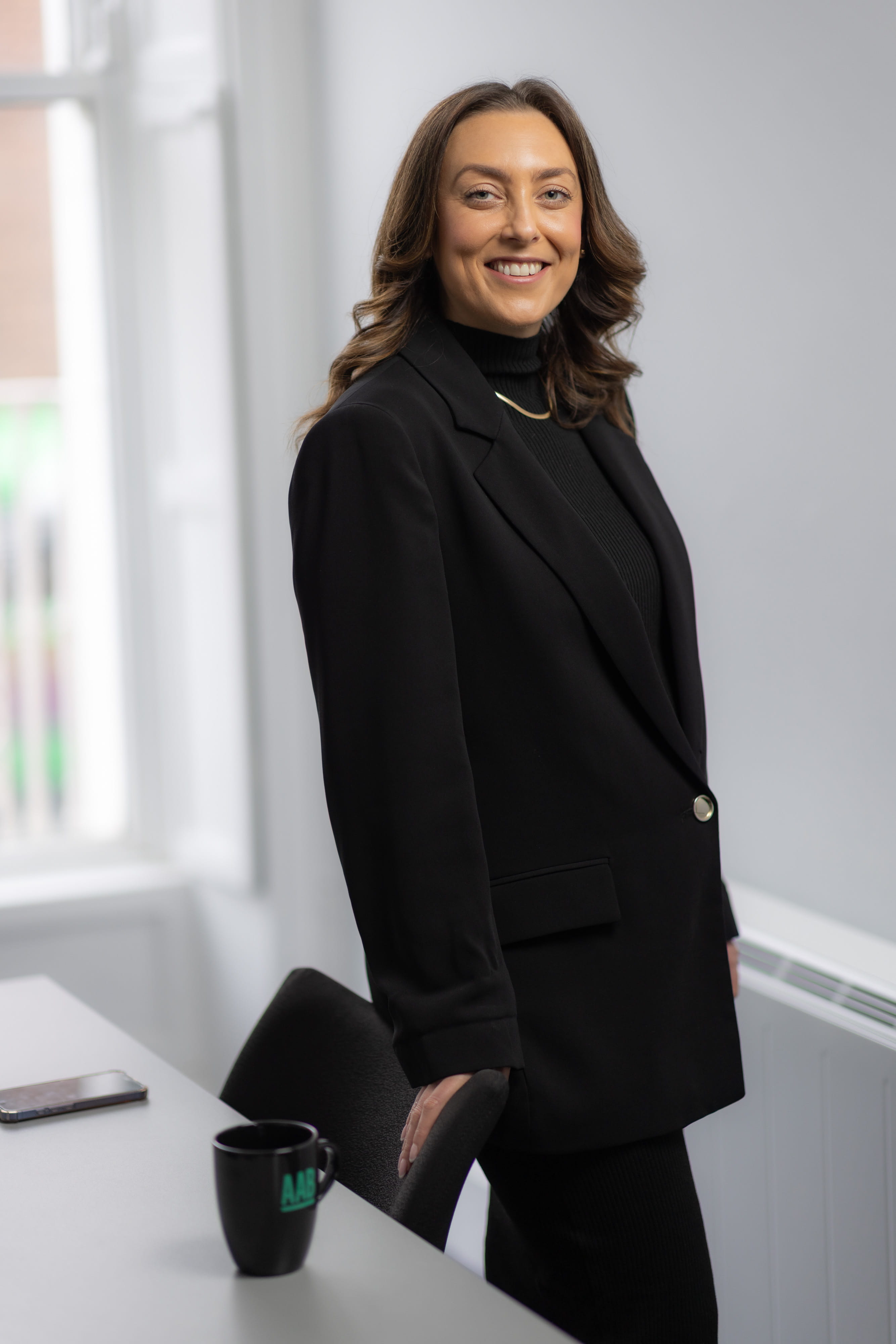 An image of Ciara McDunphy wearing a black blazer and standing next to a desk smiling.