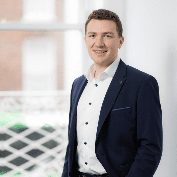 An image of Ruairi Power standing in the office wearing a black suit jacket and a white shirt. His hand is in his pocket.