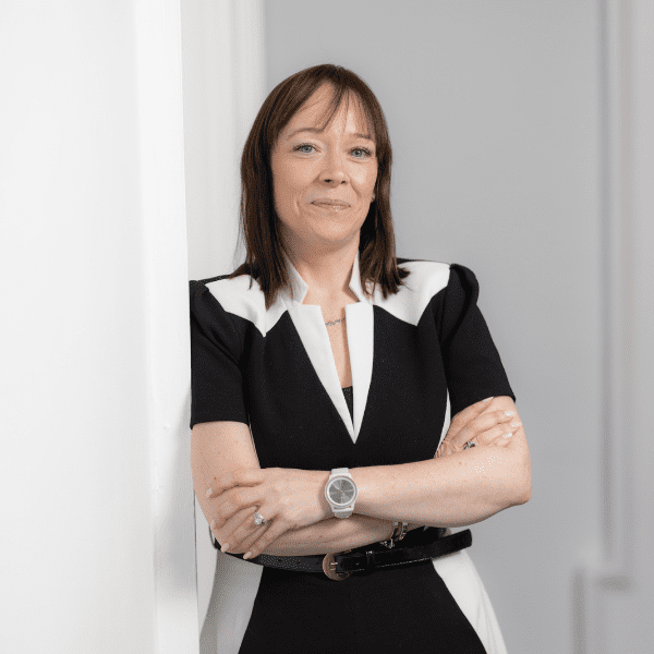 An image of Ciara O'Luanaigh wearing a black and white dress. She has brown hair and has her arms crossed smiling.