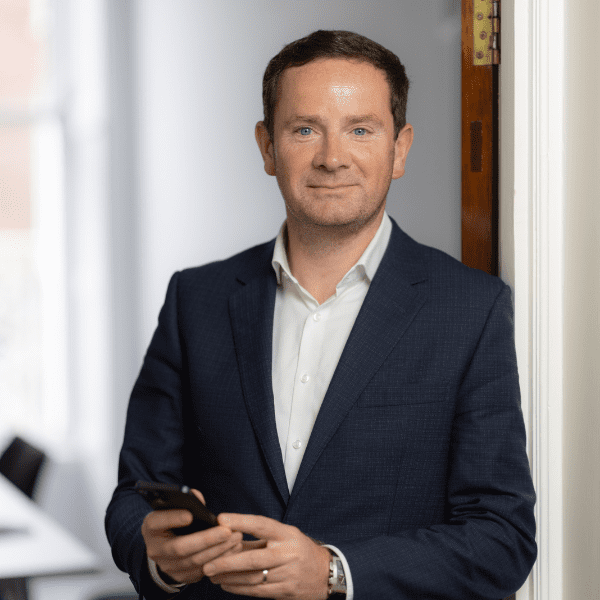 An image of Joun Conway wearing a black suit jacket with a white shirt. He is standing in the office and smiling at the camera.