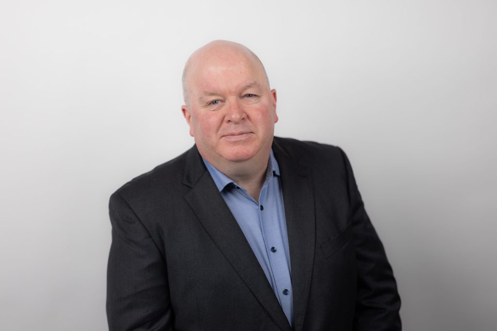 An image of Justin Brady wearing a black suit jacket with a blue shirt.