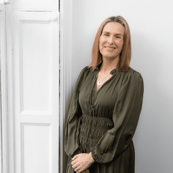 An image of Sylvia standing against a window, with her hands clasped and smiling at the camera.