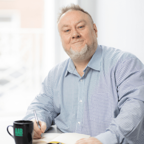 An image of Richard Maloney sitting at a desk and smiling at the camera. He is wearing a blue shirt and has an AAB mug.