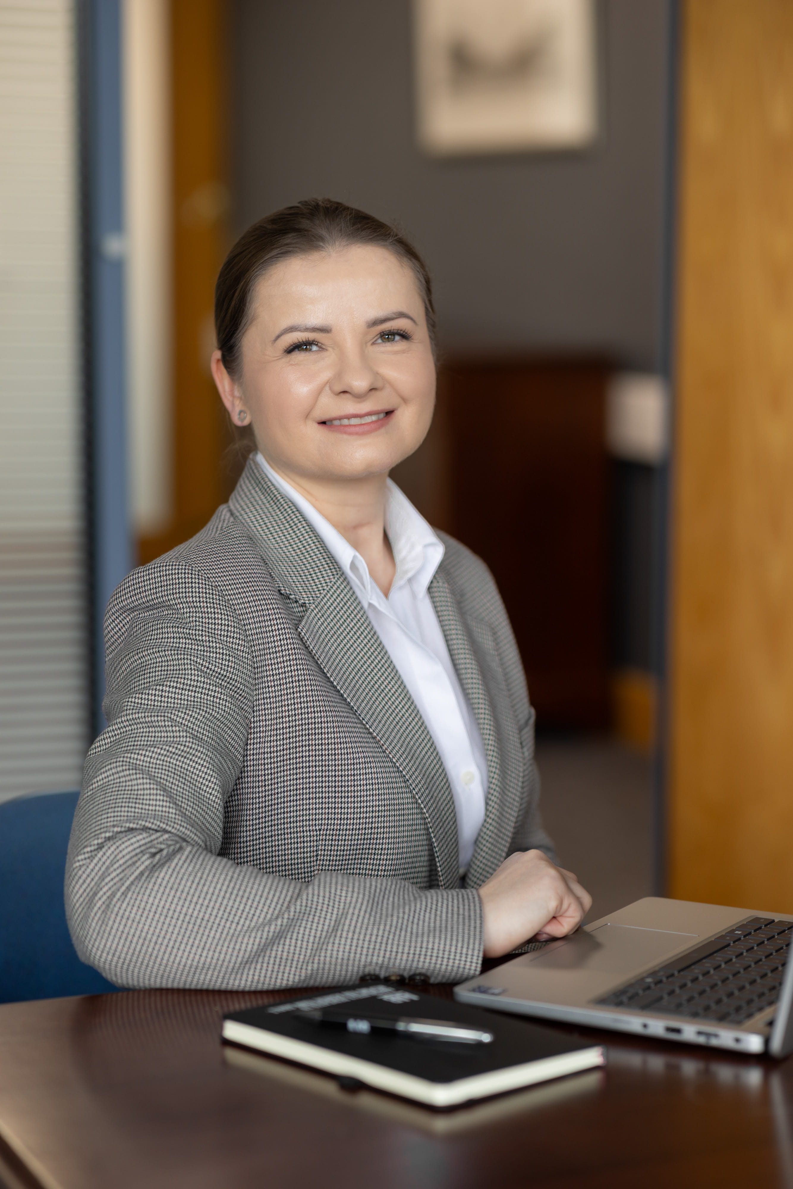 An image of Zoe Podraza Ostrega wearing a grey suit jacket and white shirt. She is sitting at the desk with her AAB notebook.