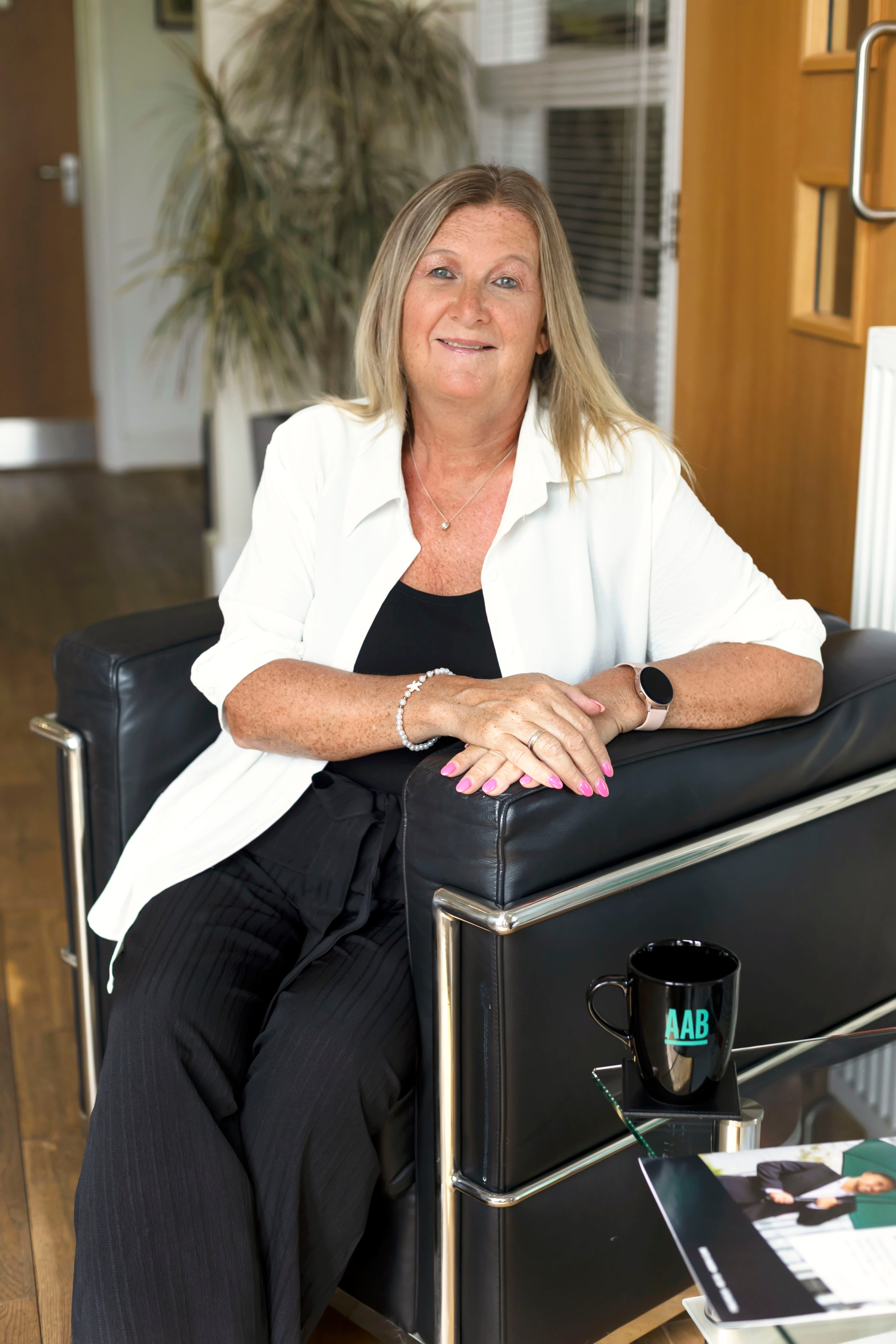An image of Karen Lane wearing a white top and black outfit. She is sitting on a chair.
