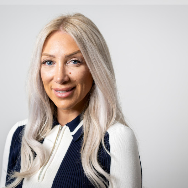 Linzi Lumbley, Director at AAB, has blonde hair and waring a white and black long sleeved dress in her headshot. The dress has white sleeves, black collar with a white line outline and white centre.