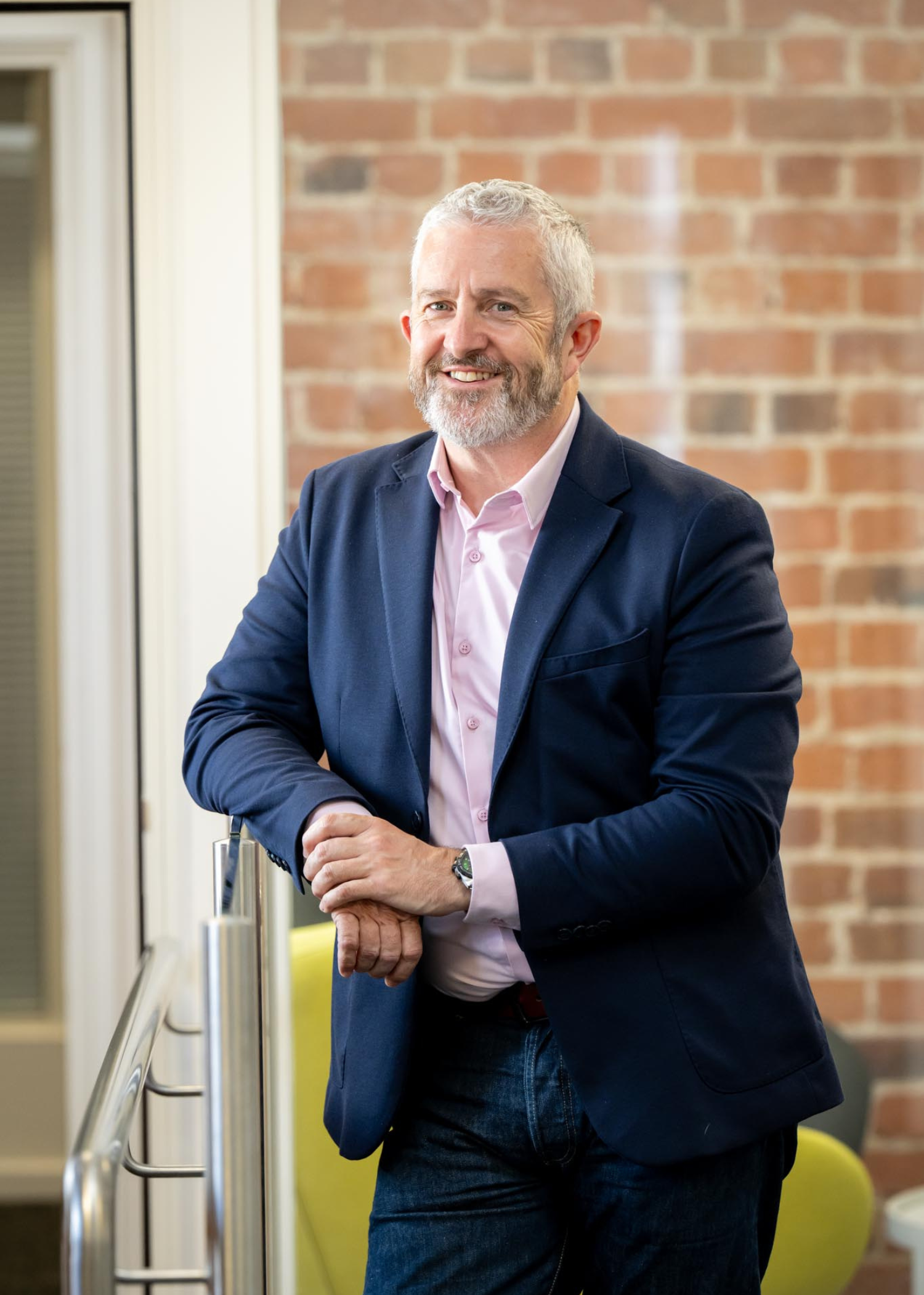 Nigel Greenaway Head of Wales and South West Region at AAB leaning against a banister with brick wall and green chairs in an office space
