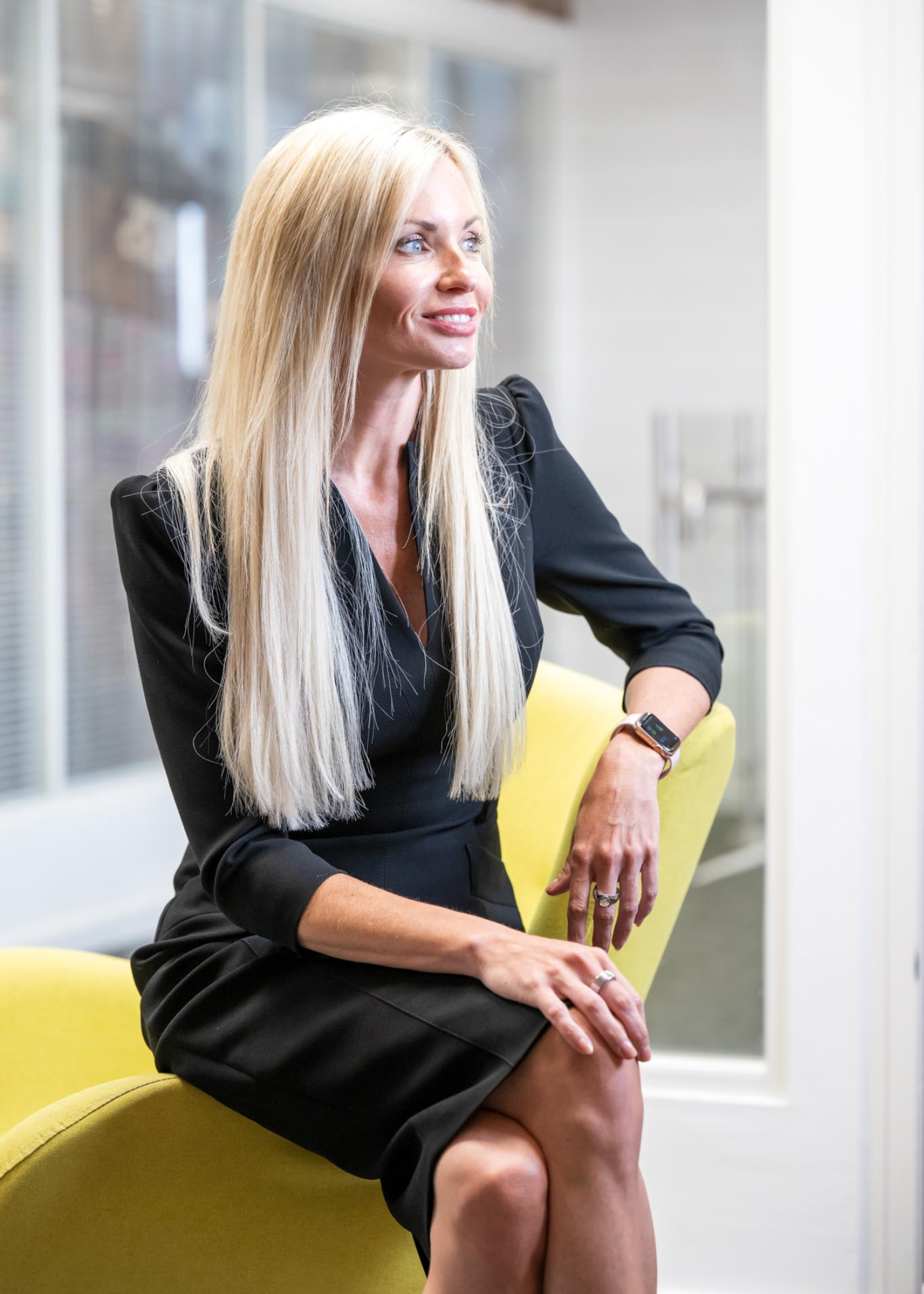 Leanne Thomas, Corporate Director at AAB, has long blonde hair and is wearing long sleeved black dress. The dress has a slight raised/padded effect on the shoulder. Leanne is sitting on the arm of a yellow chair with a glass window background