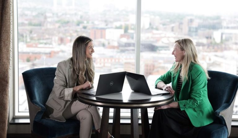 Two team members sitting by the window in the office on their laptops chatting